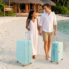 A couple strolls on a beach, each pulling a light blue luggage bag, with a tropical hut in the background.