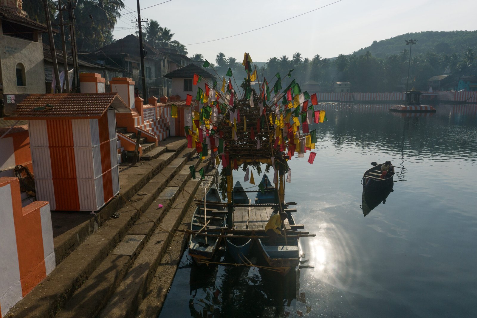 Colorful flags adorn a boat on a serene lake, with steps and buildings lining the shore under a clear sky Image for Slow Travel in South India